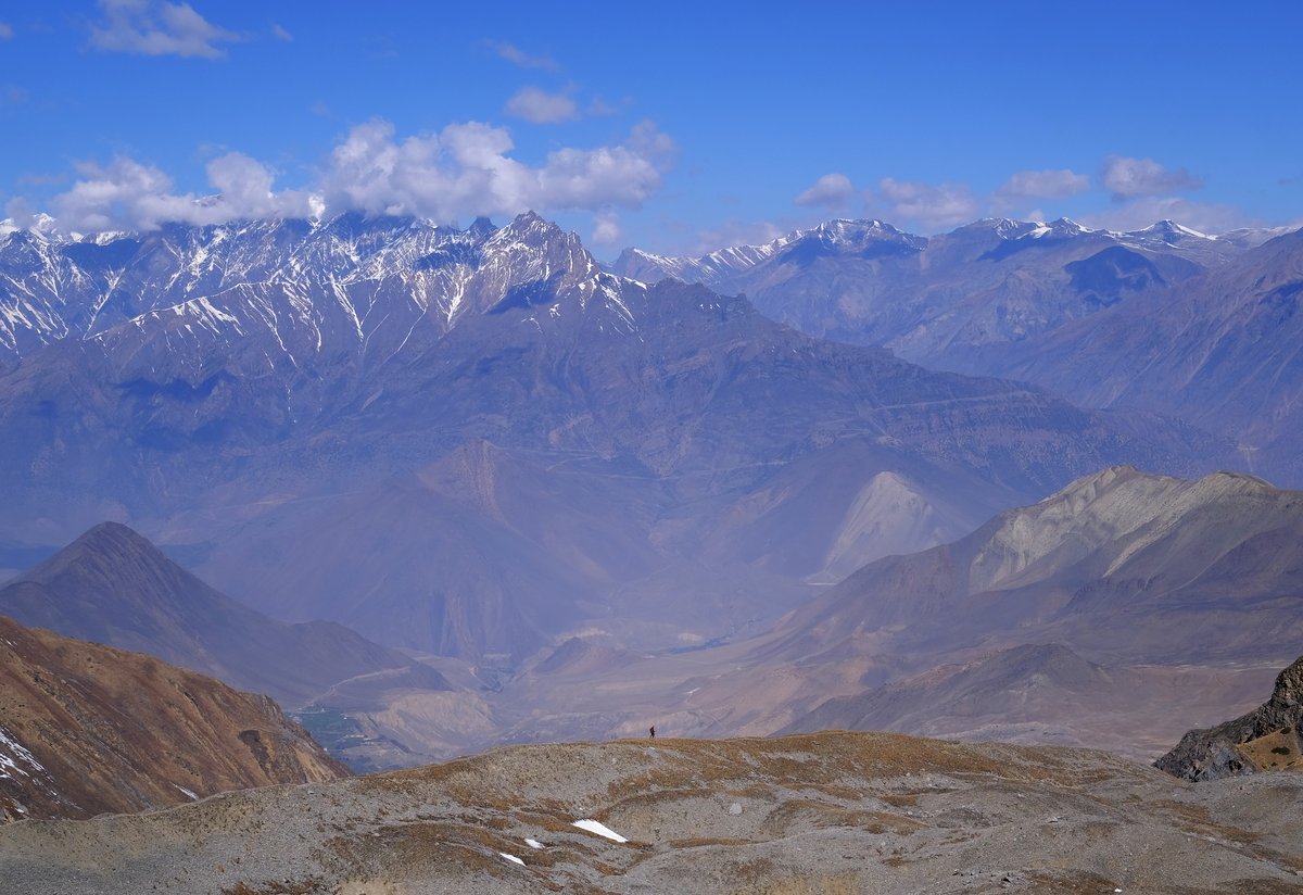 The path descending towards Mukthinath is visible at the left. A lone trekker at the center of the frame highlights the scale of the landscape